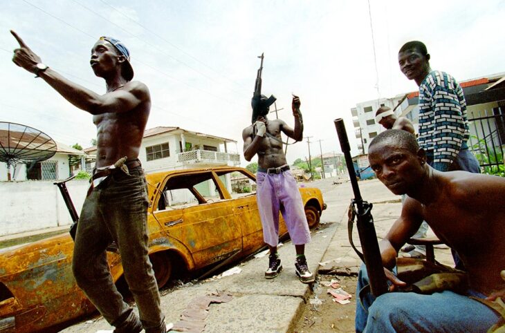 Au Liberia, des combattants du Front national patriotique du Liberia (NPFL), lourdement armés, sont rassemblés autour d'une épave de voiture. L'un d'entre eux semble s'adresser de façon virulente à une autre personne plus loin.
