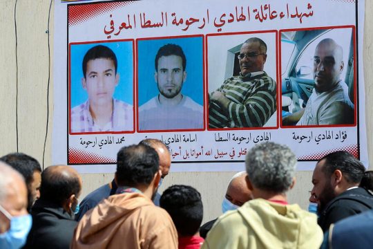 A group of people gather in the street, in Libya, in front of a poster showing 4 civilian victims of militias.