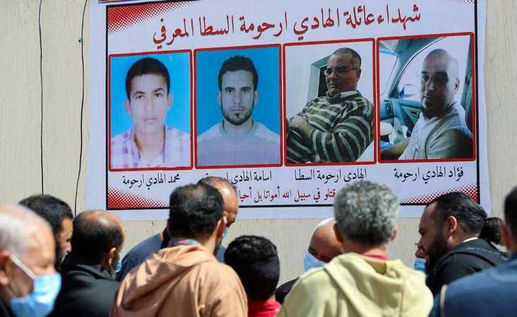 A group of people gather in the street, in Libya, in front of a poster showing 4 civilian victims of militias.