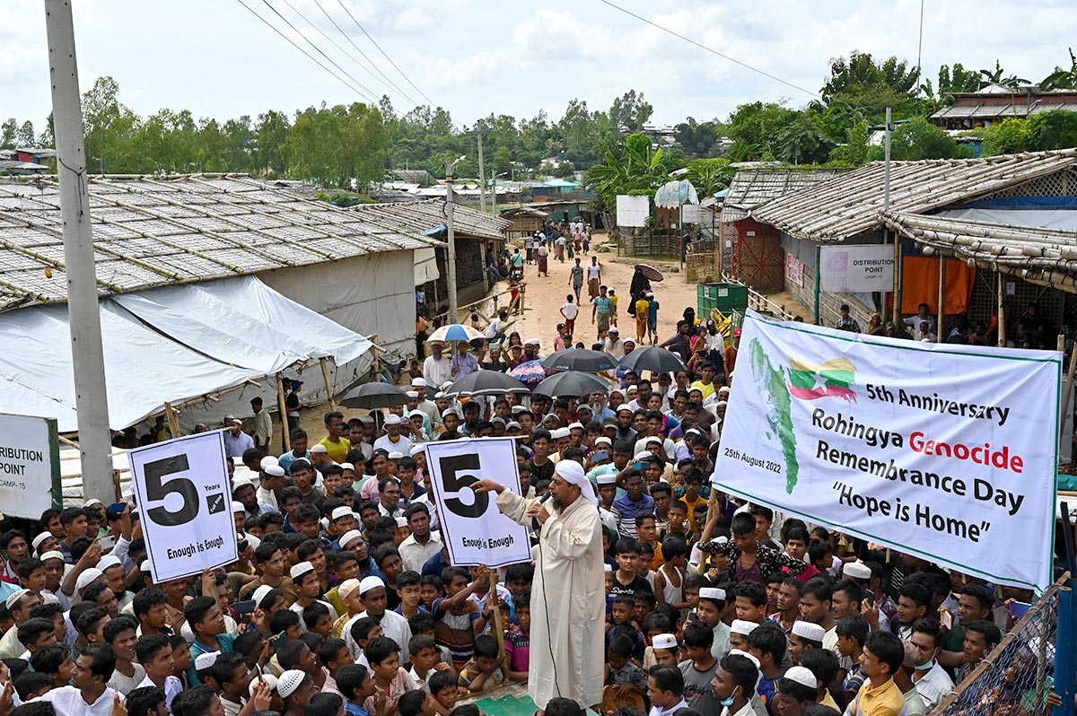 In a village street, a crowd is gathered around a person speaking at a microphone, on a platform. Banners read "5 years. Enough is enough" and "5th anniversary - Rohingya genocide remembrance day - Hope is home."
