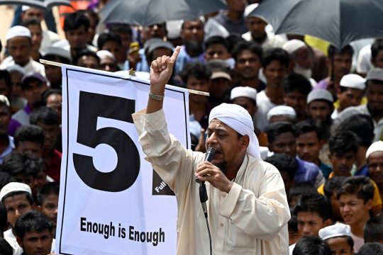 A man speaks into a microphone, on a platform, surrounded by a crowd. He raises his fist in front of a banner that reads, 