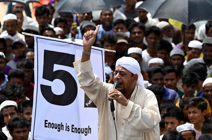 A man speaks into a microphone, on a platform, surrounded by a crowd. He raises his fist in front of a banner that reads, "5 - Enough is enough."