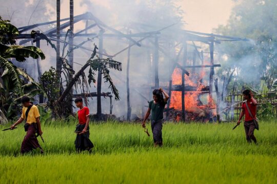 Au Myanmar, la population Rohingya a été victime en 2017 de crimes qui pourraient être reliés à une volonté de génocide, dont des destructions de terres et villages. Photo : un village brûle tandis que des hommes munis de poignards et de lance-pierres passent au premier plan.
