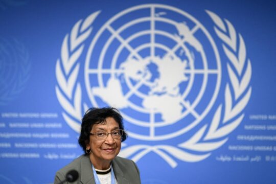 Navi Pillay, wearing glasses, a grey jacket and a UN badge on a lanyard, smiles behind a microphone at a press conference. Behind her is the large white United Nations emblem on a blue background, with “United Nations Geneva” written in several languages.