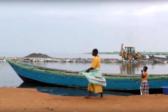 Au premier plan, un bateau et des habitants. En arrière-plan, une pelleteuse.