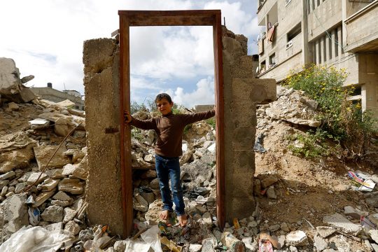 Palestinian child standing in front of a gate without walls around it, in the middle of the ruins