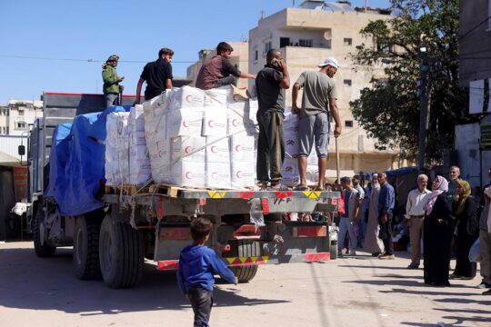 Un camion rempli de paquets, sur lesquels sont postés des hommes, est arrêté devant un groupe de personnes attendant calmement.