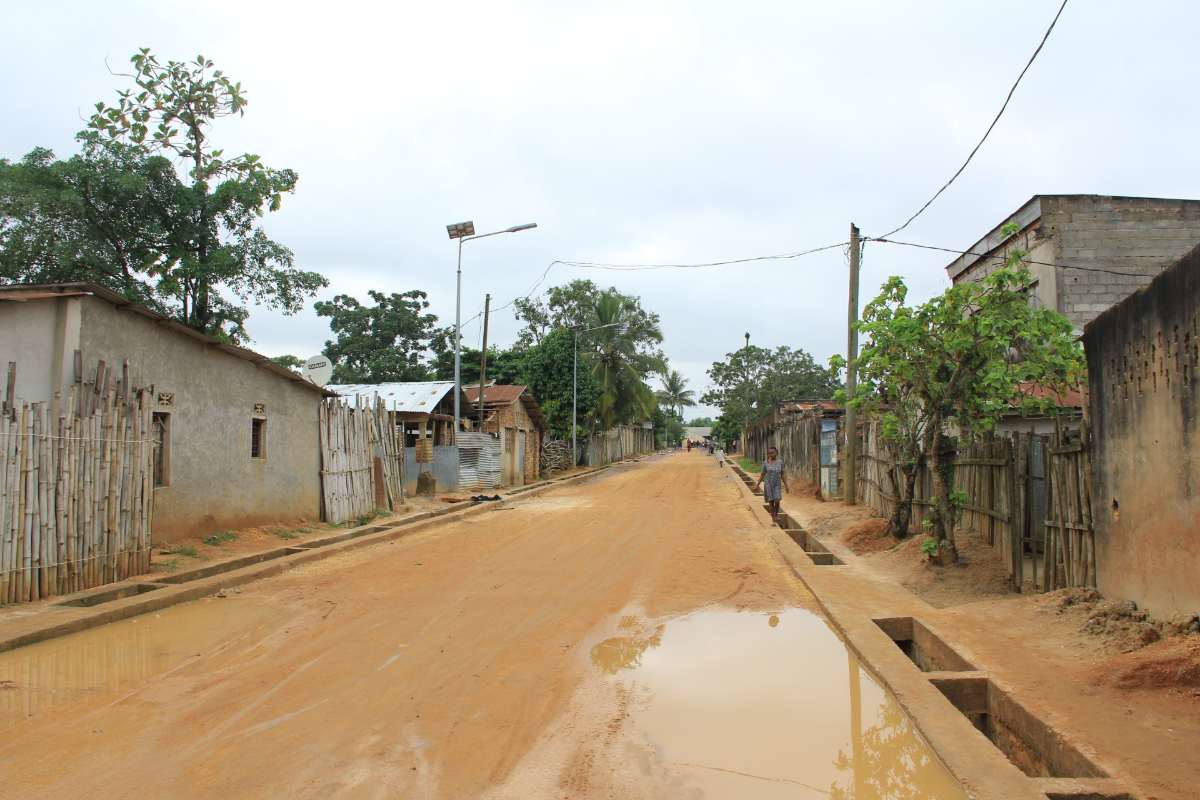 Une large rue de terre rouge du quartier des musiciens à Kisangani, bordée de maisons basses et de clôtures en bambou ; une flaque d'eau au premier plan et une personne marchant au loin sous un ciel couvert.
