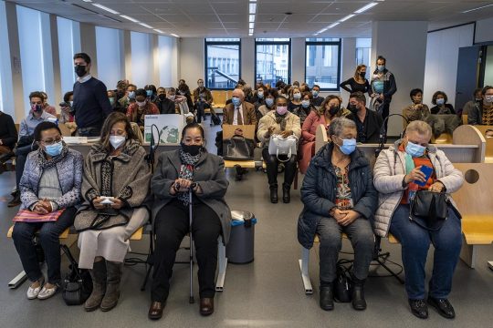 Several people are sitting in a waiting room with 5 women in the front row.