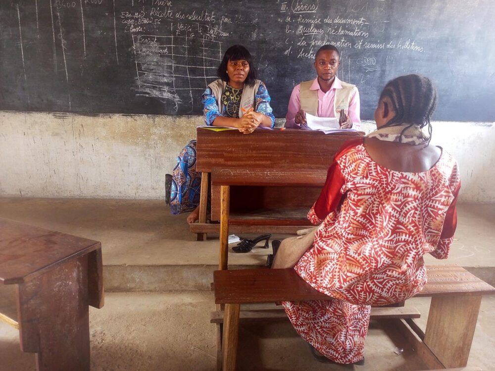 On Saturday 4 July 2026, a team of investigators from the Provincial Truth, Justice and Reconciliation Commission takes statements from victims on the premises of Mukoleshi School, in the commune of Nganza (Kasai-Central Province, Democratic Republic of the Congo). Photo: © Caleb Kazadi Seated on a school bench, a woman is seen from behind, facing two people (seated in the teacher’s chair) who appear to be listening to her, during the victims’ hearings before the Kasai Truth Commission in the Democratic Republic of the Congo (CVPJR).