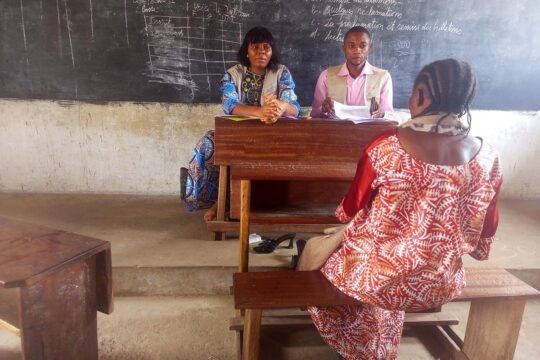 Seated on a school bench, a woman is seen from behind, facing two people (seated in the teacher’s chair) who appear to be listening to her, during the victims’ hearings before the Kasai Truth Commission in the Democratic Republic of the Congo (CVPJR).