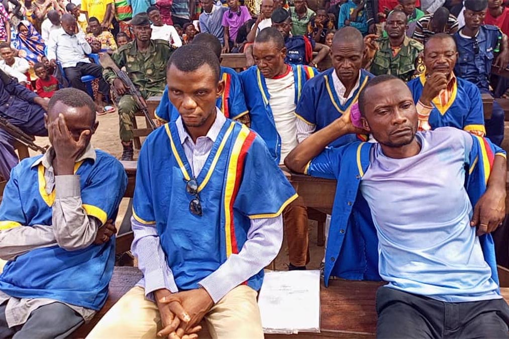 The seven defendants in the Kamuina Nspau trial in Kasai, DRC, wear colorful uniforms and sit on wooden benches in the open air.
