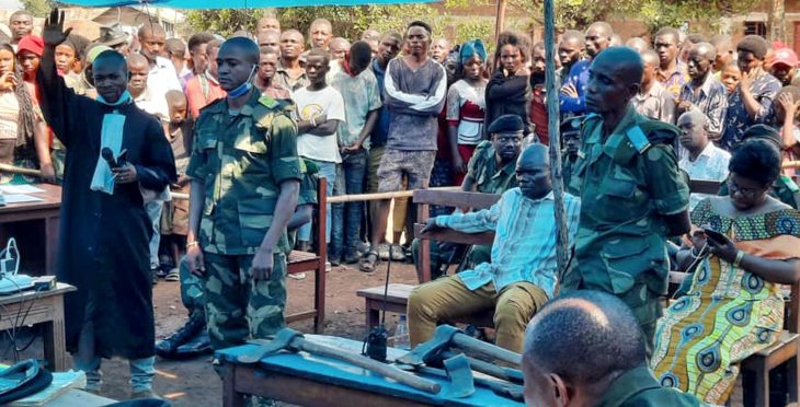 An outdoor trial with lawyers, military, civilians and the public standing around