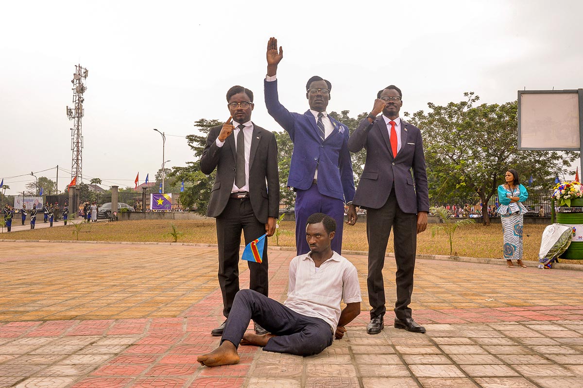 4 acteurs imitent Patrice Lumumba. 3 debouts (en costume, fiers, mains et poings levés), en costume, tandis que le 4ème est assis par terre, pieds nus et les mains dans le dos.