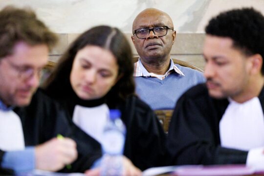 Procès en Belgique d'Emmanuel Nkunduwimye, accusé de génocide au Rwanda. Photo : Nkunduwimye fixe l'objectif derrière ses avocats qui discutent.