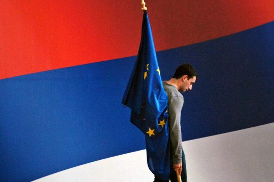 Un homme est debout sur une estrade avec un grand drapeau de la Serbie en arrière-plan. Il tient un petit drapeau de l'Union européenne.