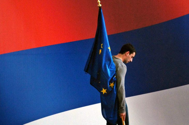 Un homme est debout sur une estrade avec un grand drapeau de la Serbie en arrière-plan. Il tient un petit drapeau de l'Union européenne.
