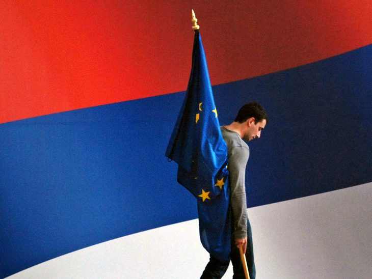 Un homme est debout sur une estrade avec un grand drapeau de la Serbie en arrière-plan. Il tient un petit drapeau de l'Union européenne.