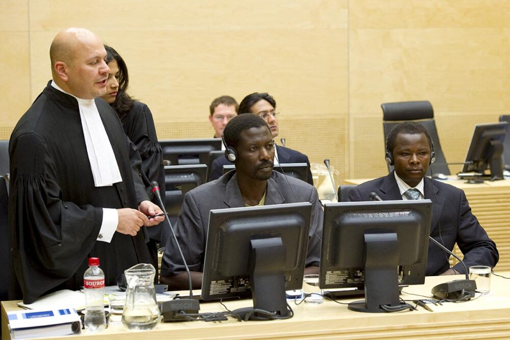 In the International Criminal Court (ICC), two men accused of war crimes in Darfur (Sudan) – Abdallah Banda Abakaer Nourain and Saleh Mohammed Jerbo Jamus – are seated next to Karim Khan (a former defence lawyer who has become a prosecutor at the ICC), who is standing in his judicial robes.