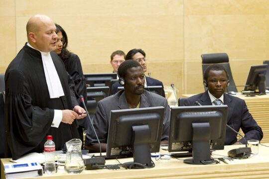 In the International Criminal Court (ICC), two men accused of war crimes in Darfur (Sudan) – Abdallah Banda Abakaer Nourain and Saleh Mohammed Jerbo Jamus – are seated next to Karim Khan (a former defence lawyer who has become a prosecutor at the ICC), who is standing in his judicial robes.