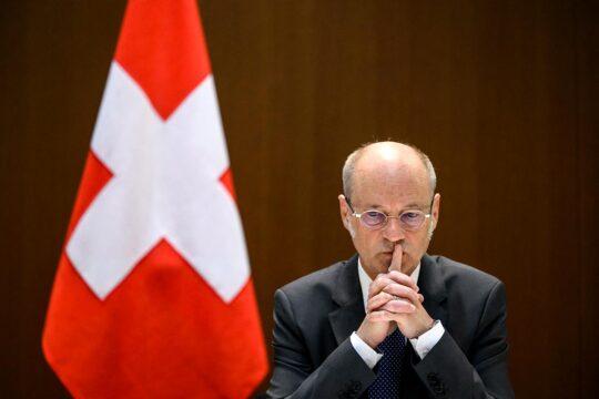 La justice internationale suisse (sous le principe de la compétence universelle) s'enlise dans des lenteurs et des suspicions d'ingérences politiques. Photo : Stefan Blattler, procureur général, en pleine réflexion, à côté d'un drapeau suisse.