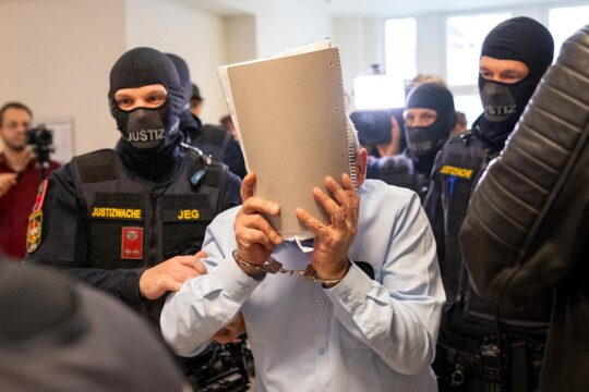 Trial in Austria of a former Syrian general for crimes committed in Syria. Photo: a man covers his face with a file as he is escorted by hooded police officers.