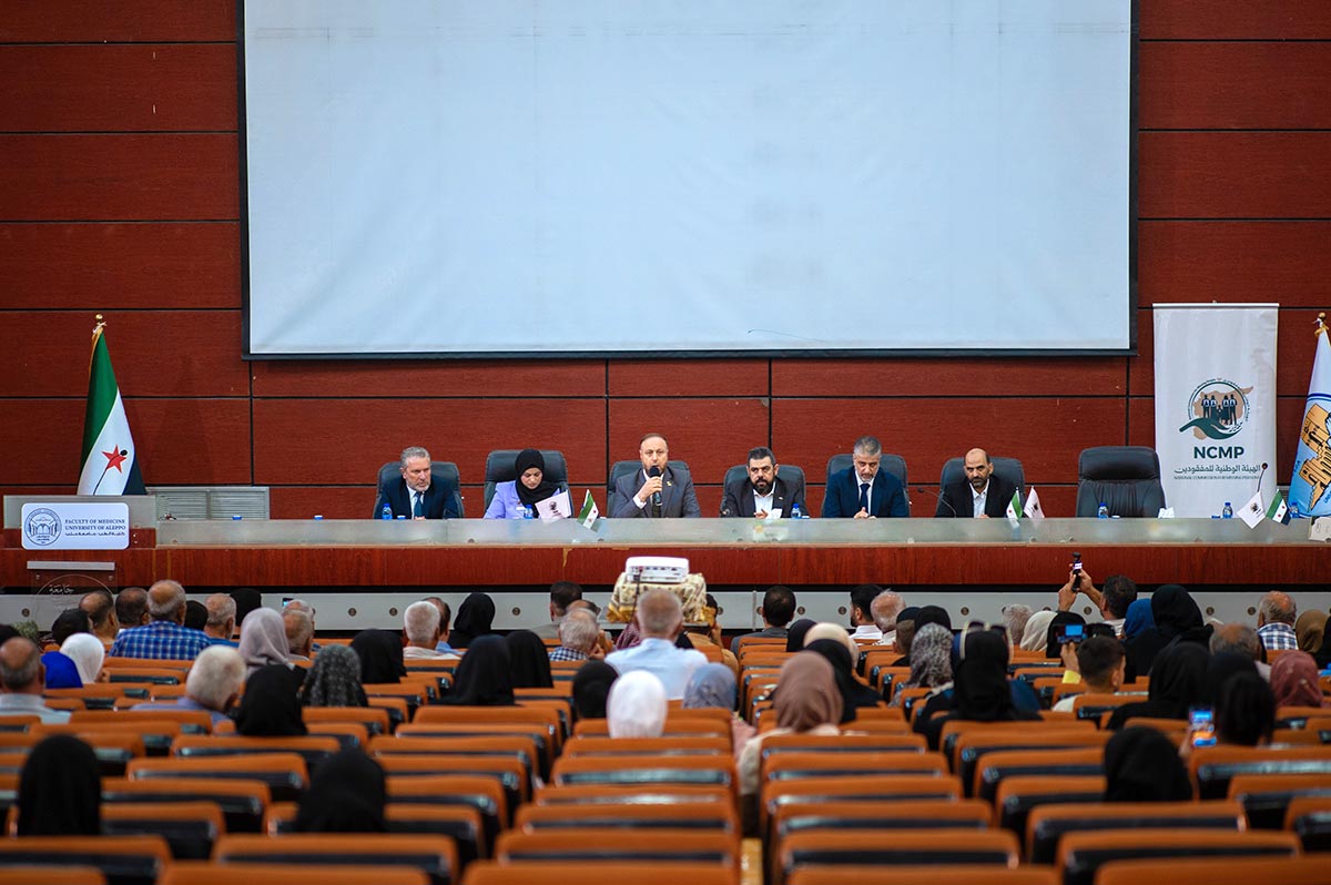 Réunion de la Commission nationale pour les personnes disparues (Syrie) dans un amphithéâtre de la faculté d’Alep. Photo : des représentants de la commission et d’autres personnalités politiques syriennes sont assis sur une estrade face aux proches des victimes, dans les gradins.