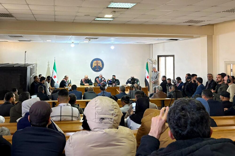 A Syrian court based in Aleppo is holding a public trial of the alleged perpetrators of crimes committed in March 2025 in Syria. Photo: a packed courtroom surrounded by journalists, with an iron cage on one side.