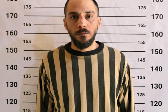 Amjad Youssef, le jour de son arrestation et de sa mise en détention à Damas (Syrie). Il est accusé d'avoir participé au massacre de Tadamon. Photo : Youssef, en costume de prisonnier rayé, pose devant un mur affichant une règle (photo officielle de prisonnier).