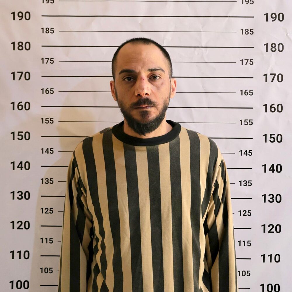Amjad Youssef, le jour de son arrestation et de sa mise en détention à Damas, le 24 avril 2026. Amjad Youssef, le jour de son arrestation et de sa mise en détention à Damas (Syrie). Il est accusé d'avoir participé au massacre de Tadamon. Photo : Youssef, en costume de prisonnier rayé, pose devant un mur affichant une règle (photo officielle de prisonnier).