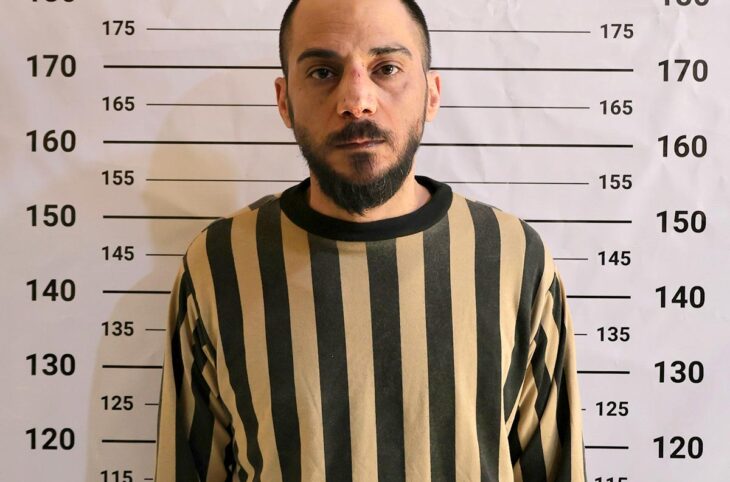 Amjad Youssef, le jour de son arrestation et de sa mise en détention à Damas (Syrie). Il est accusé d'avoir participé au massacre de Tadamon. Photo : Youssef, en costume de prisonnier rayé, pose devant un mur affichant une règle (photo officielle de prisonnier).