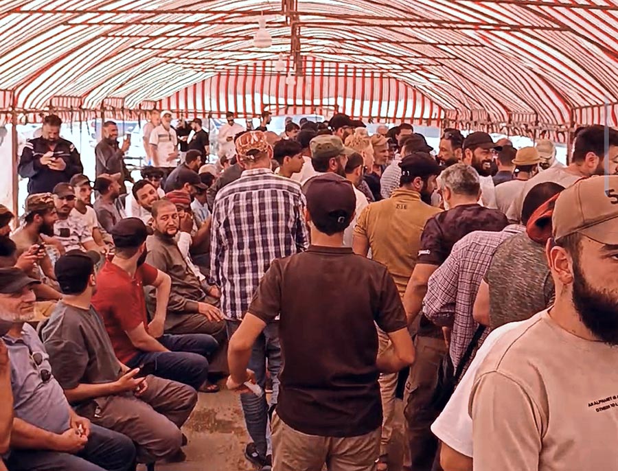 A Deir ez-Zor (Syrie) une manifestation prend la forme d'un sit-in ouvert baptisé “Sit-in de la dignité”. Une tente est installée. Photo : de nombreux hommes (certains assis, d'autres debout) se tiennent sous une tente rayée blanche et rouge.