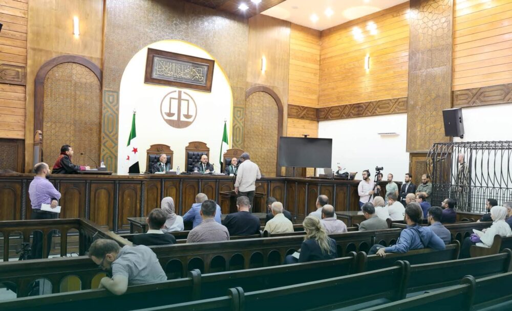 A hearing before the Fourth Criminal Court in Damascus in July 2026 in the trial of Wasim al-Assad. In August, the court handed down several verdicts in cases involving serious crimes committed under the Assad family’s regime. Photo : © Syrian Ministry of Justice In Syria, people are on trial for their involvement in crimes committed under Bashar al-Assad’s regime. Photo: The trial of Wasim al-Assad, standing in a cage at the side of a relatively empty courtroom.