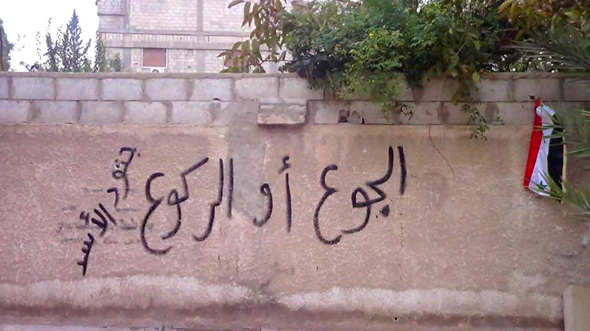 Famine et crimes de guerre en Syrie - Photo : un graffiti sur un mur clame en arabe « À genoux ou affamés ! ». Sur le côté, un drapeau syrien.