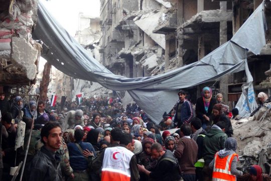 Dans une rue dont les bâtiments sont complètement dévastés (quartier de Yarmouk à Damas, en Syrie), une distribution d'aide alimentaire est organisée auprès de nombreux réfugiés