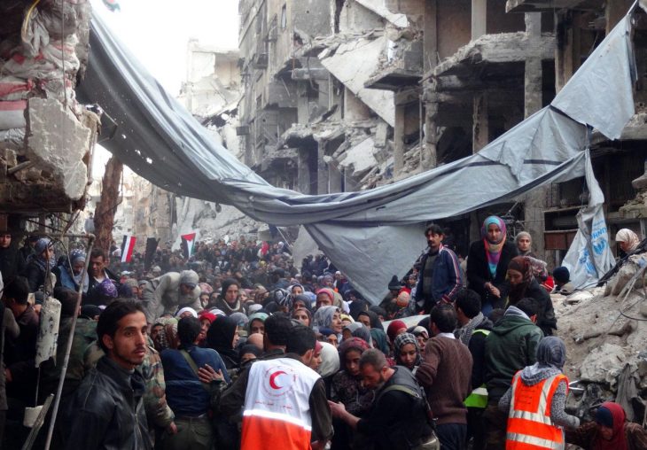 Dans une rue dont les bâtiments sont complètement dévastés (quartier de Yarmouk à Damas, en Syrie), une distribution d'aide alimentaire est organisée auprès de nombreux réfugiés
