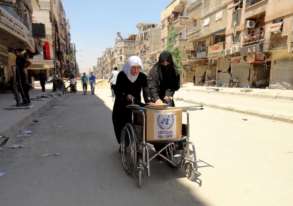 Famine et crimes de guerre en Syrie : un procès à Coblence (Allemagne) tente de faire le lien. Photo : deux femmes voilées poussent, dans une rue détruite de Damas, un fauteuil roulant sur lequel est disposé un colis humanitaire de l'UNRWA.