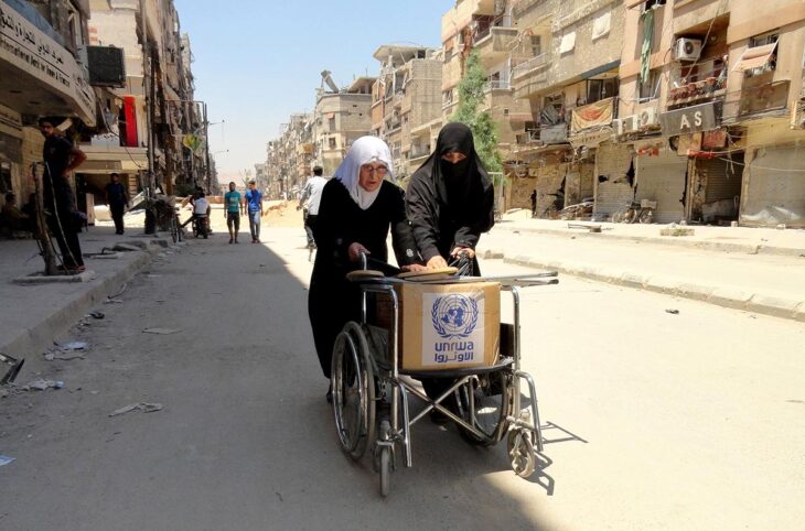 Famine et crimes de guerre en Syrie : un procès à Coblence (Allemagne) tente de faire le lien. Photo : deux femmes voilées poussent, dans une rue détruite de Damas, un fauteuil roulant sur lequel est disposé un colis humanitaire de l'UNRWA.