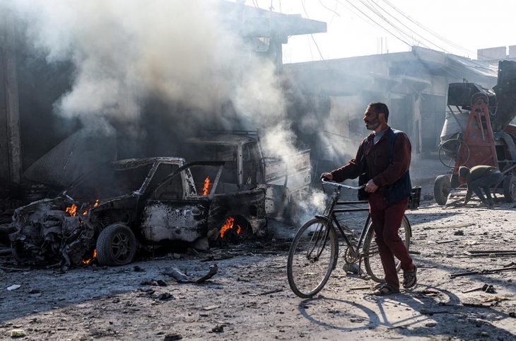 Syrian civilian riding his bike through the street near the remains of a car bomb explosion