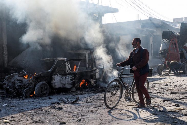 Syrian civilian riding his bike through the street near the remains of a car bomb explosion