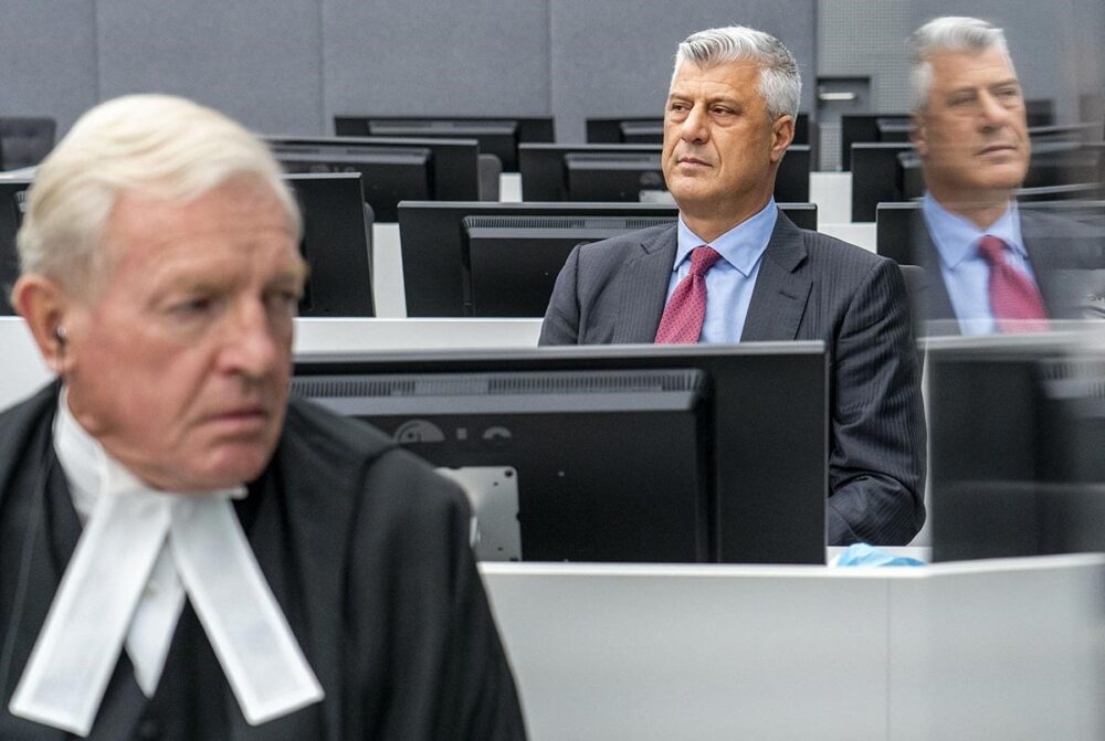 L'ancien président du Kosovo, Hashim Thaçi, devant les Chambres spécialisées pour le Kosovo, à La Haye, en novembre 2020. Après six ans de procédure, il a été condamné à 25 ans de prison pour crimes de guerre. Photo : © Jerry Lampen / ANP / AFP Hashim Thaçi, ancien président du Kosovo, assis dans le box des accusés des Chambres spécialisées pour le Kosovo à La Haye, en costume sombre, chemise bleue et cravate rouge, le regard fixe. Au premier plan, flou, un avocat en robe et rabat blanc. Le reflet de Thaçi apparaît dans une vitre à droite.