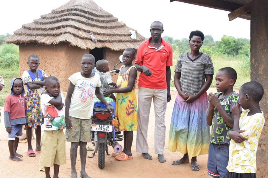 Geoffrey Byakagaba poses in the center of his family in their village of Kasenyi, Uganda. In the background is a modest house with mud walls and a thatched roof.