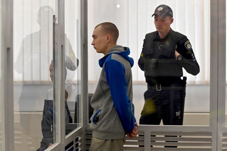 Vadim Shishimarin, a Russian soldier accused of war crimes, stands in the dock during his trial in Ukraine. A police officer is standing behind him.