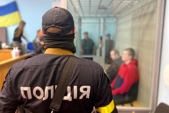 Un policier ukrainien de dos. En arrière-plan, les 2 soldats russes accusés, Aleksandr Bobykin et Aleksandr Ivanov, dans le box, lors de leur procès en Ukraine.