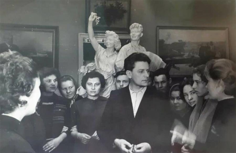 L’artiste Albin Havdzynskyi photographié en noir et blanc dans une galerie d'art (peintures et sculptures en arrière-plan), entouré de femmes et d'hommes, avec qui il semble discuter.