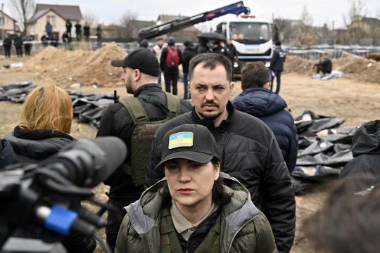 Iryna Venediktova and a man face the press. In the background, corpses wrapped in body bags, men and a crane.