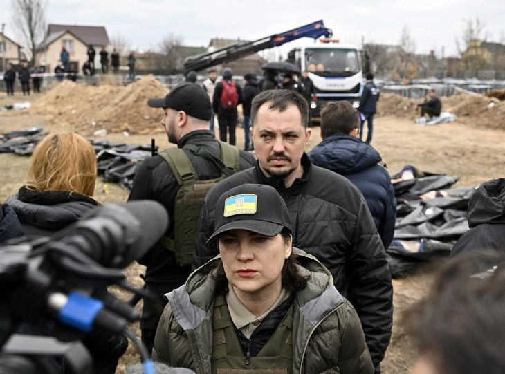 Iryna Venediktova and a man face the press. In the background, corpses wrapped in body bags, men and a crane.