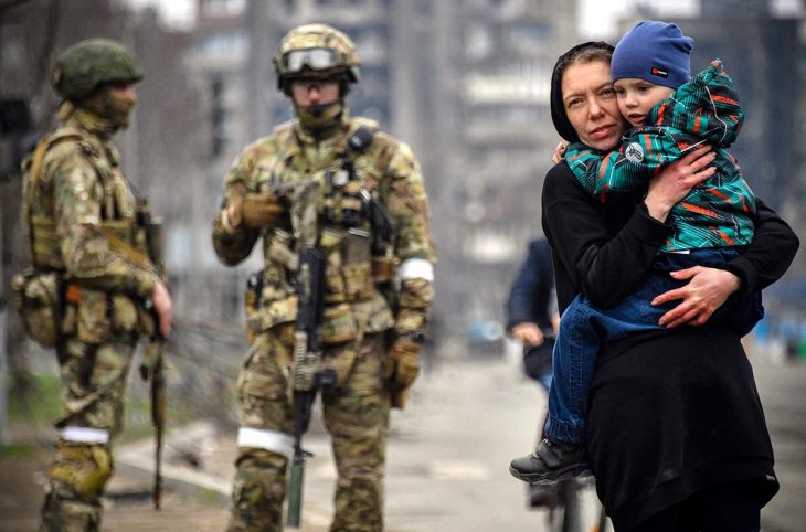 In Marioupol, Ukraine, a woman carries her child in her arms in the street. Behind her, Russian soldiers are patrolling. In the background: destroyed civilian buildings.