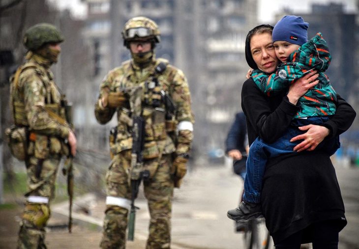 In Marioupol, Ukraine, a woman carries her child in her arms in the street. Behind her, Russian soldiers are patrolling. In the background: destroyed civilian buildings.