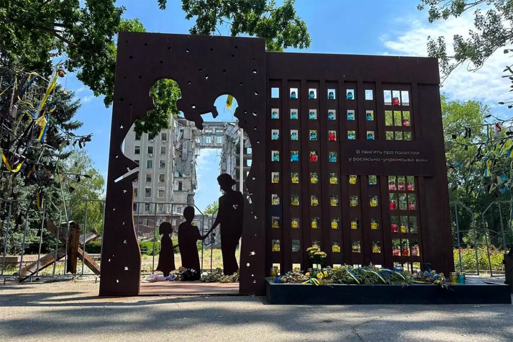 Monuments en mémoire des crimes de guerre en Ukraine. Photo : un monumemt dédié aux victimes de crimes de guerre représente une famille disparue qu'on voit au travers d'un mur percé. En arrière-plan, un bâtiment de plusieurs étages détruit.